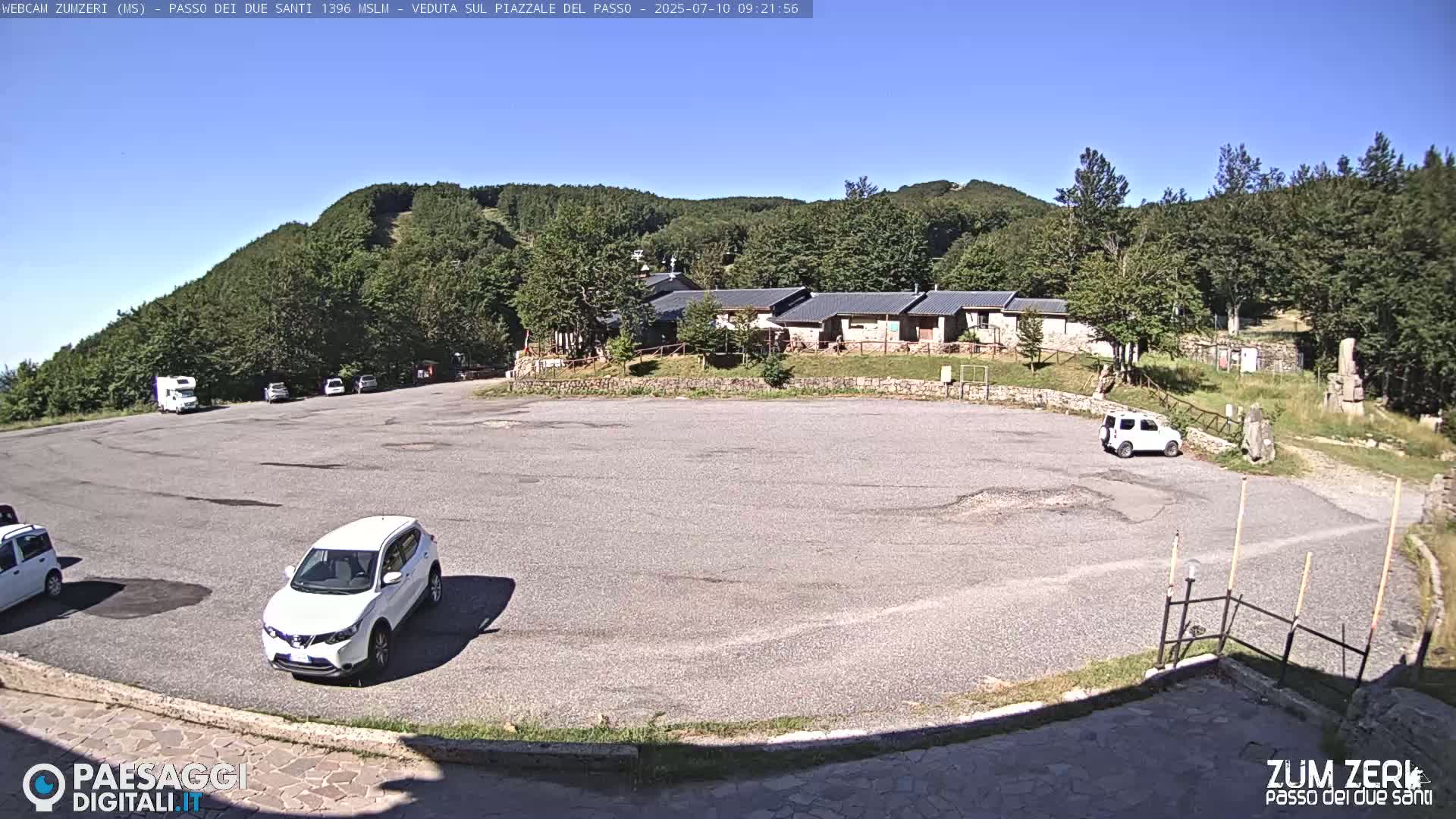 A mostly empty asphalt parking lot sits in front of a building complex nestled among green, tree-covered hills under a clear blue sky.