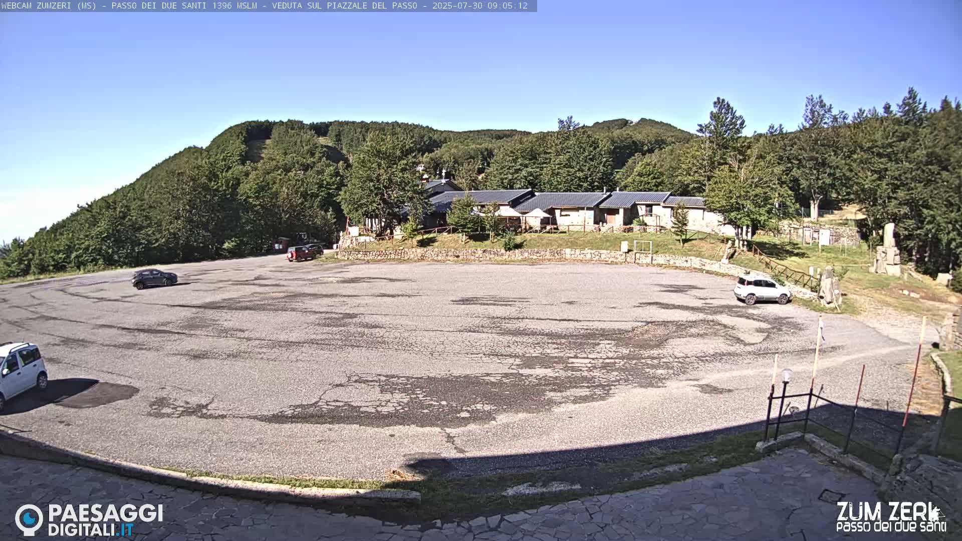 A mostly empty asphalt parking lot sits below a cluster of buildings nestled against a verdant hillside under a clear blue sky.