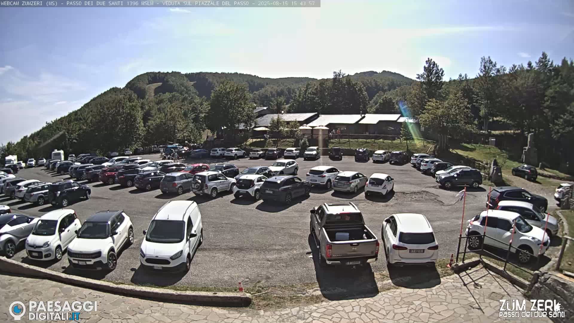 A mostly empty asphalt parking lot sits below a cluster of buildings nestled against a verdant hillside under a clear blue sky.