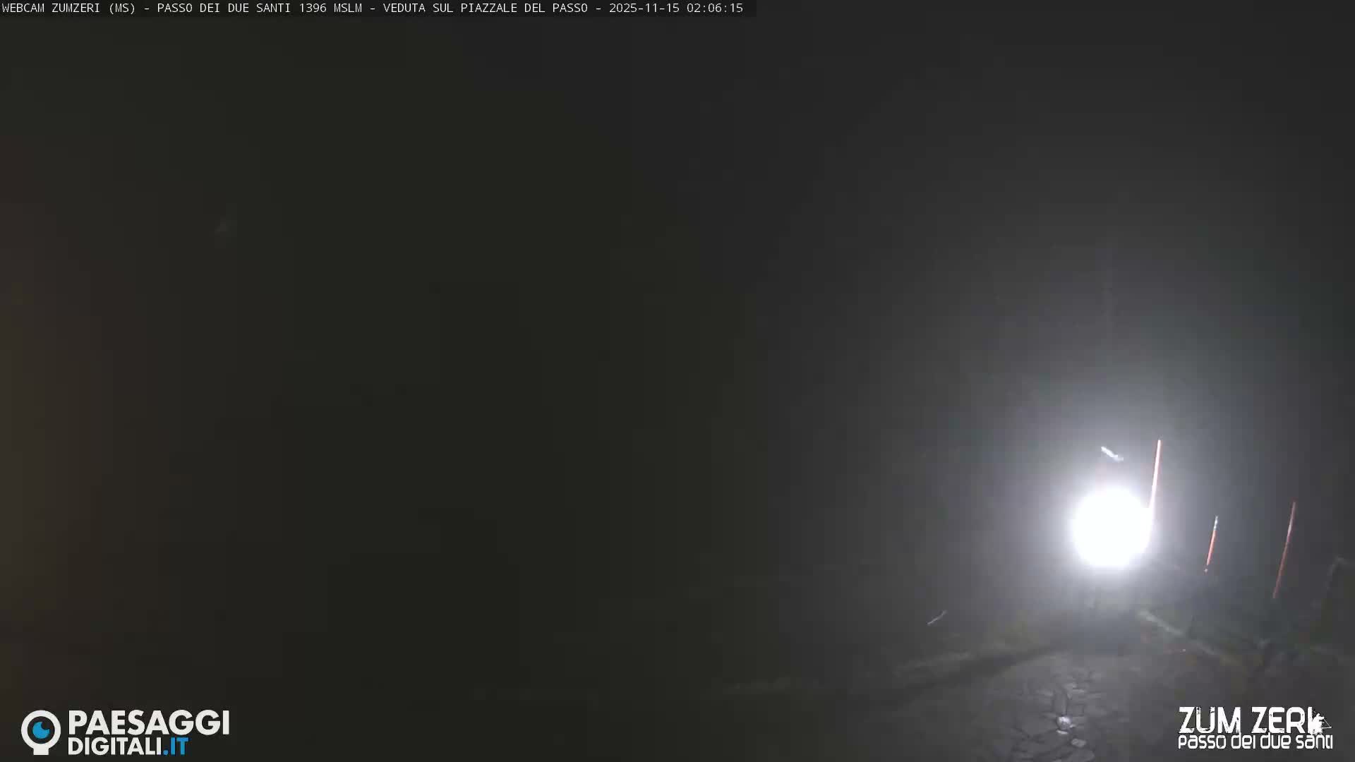 The image displays a dark, foggy night scene outdoors, illuminated by a bright, diffused light source in the lower right, with faint vertical markers visible on the right.