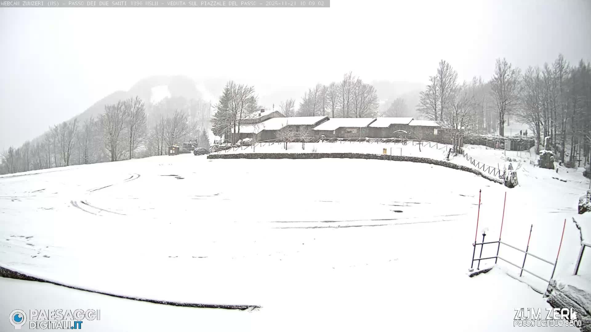 A winter scene under an overcast sky showcases a vast snow-covered field with tire tracks, a cluster of buildings with snow-laden roofs, and surrounding hills dotted with bare, snow-dusted trees.