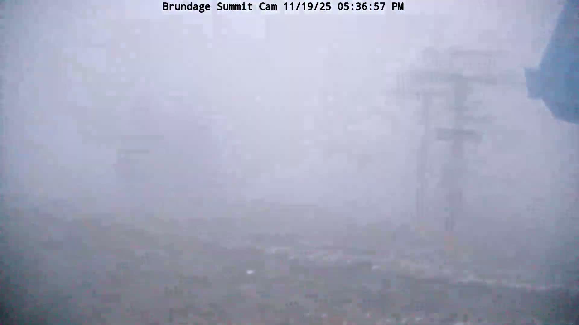 A mountain ski slope is heavily obscured by thick fog and falling snow, with faint outlines of ski lift towers barely visible through the very low visibility.