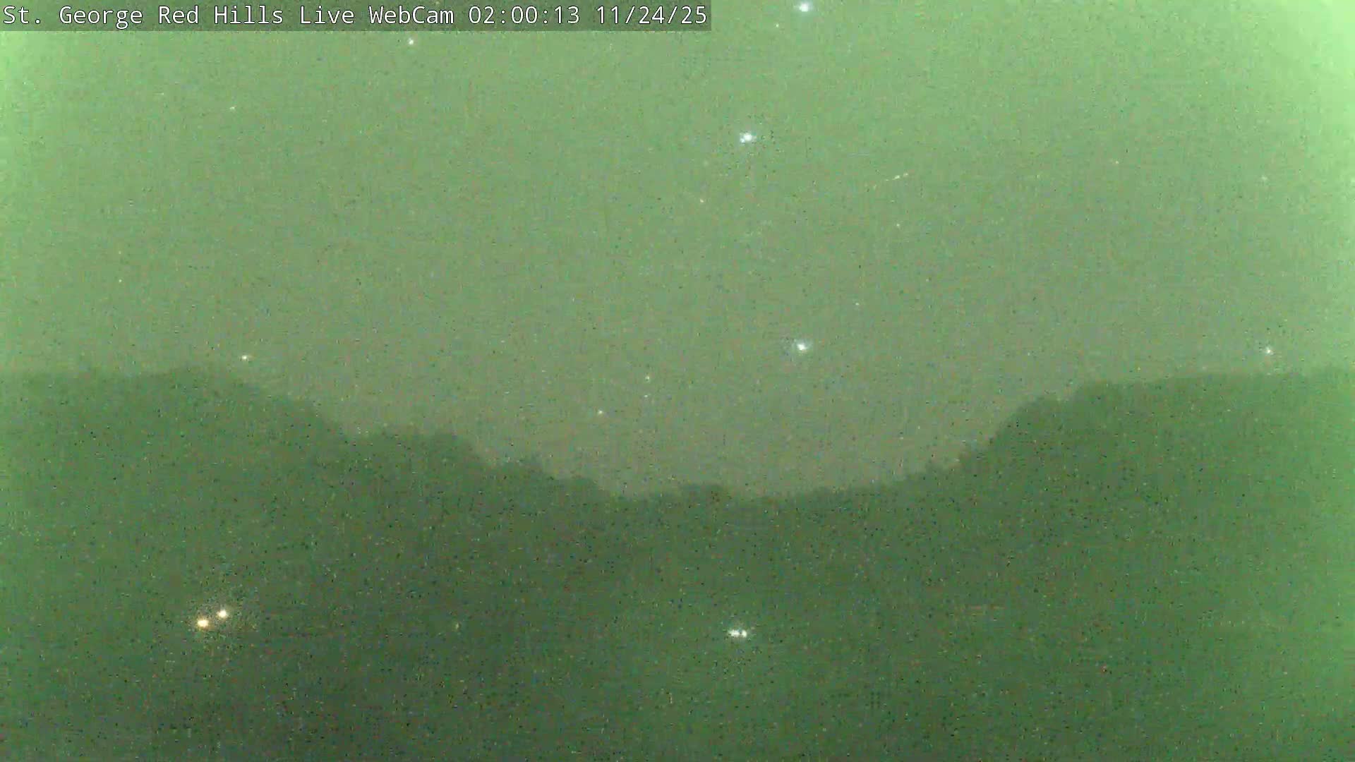 A very grainy, green-tinted nighttime view reveals dark, undulating hills against a lighter sky dotted with distinct bright points of light, suggesting clear conditions.