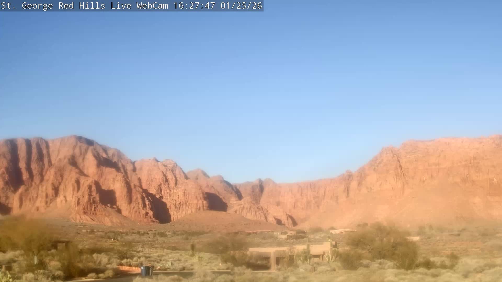 The image shows a wide view of towering red rock mountains under a clear blue sky, with desert vegetation and a few low-rise buildings in the foreground on a sunny day.