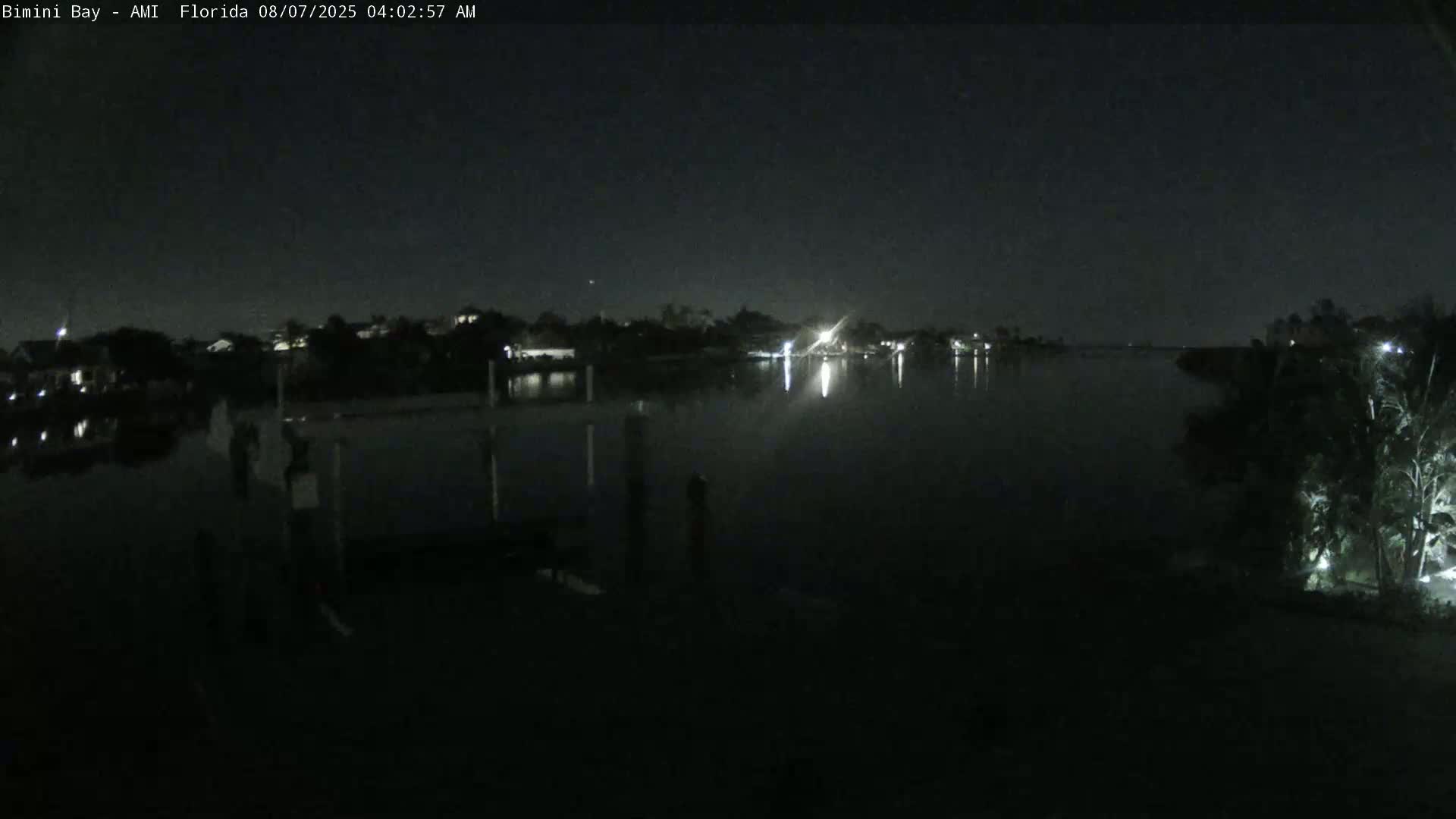 A nighttime view of a calm body of water reflecting the lights of houses across the shore under a dark, clear sky.