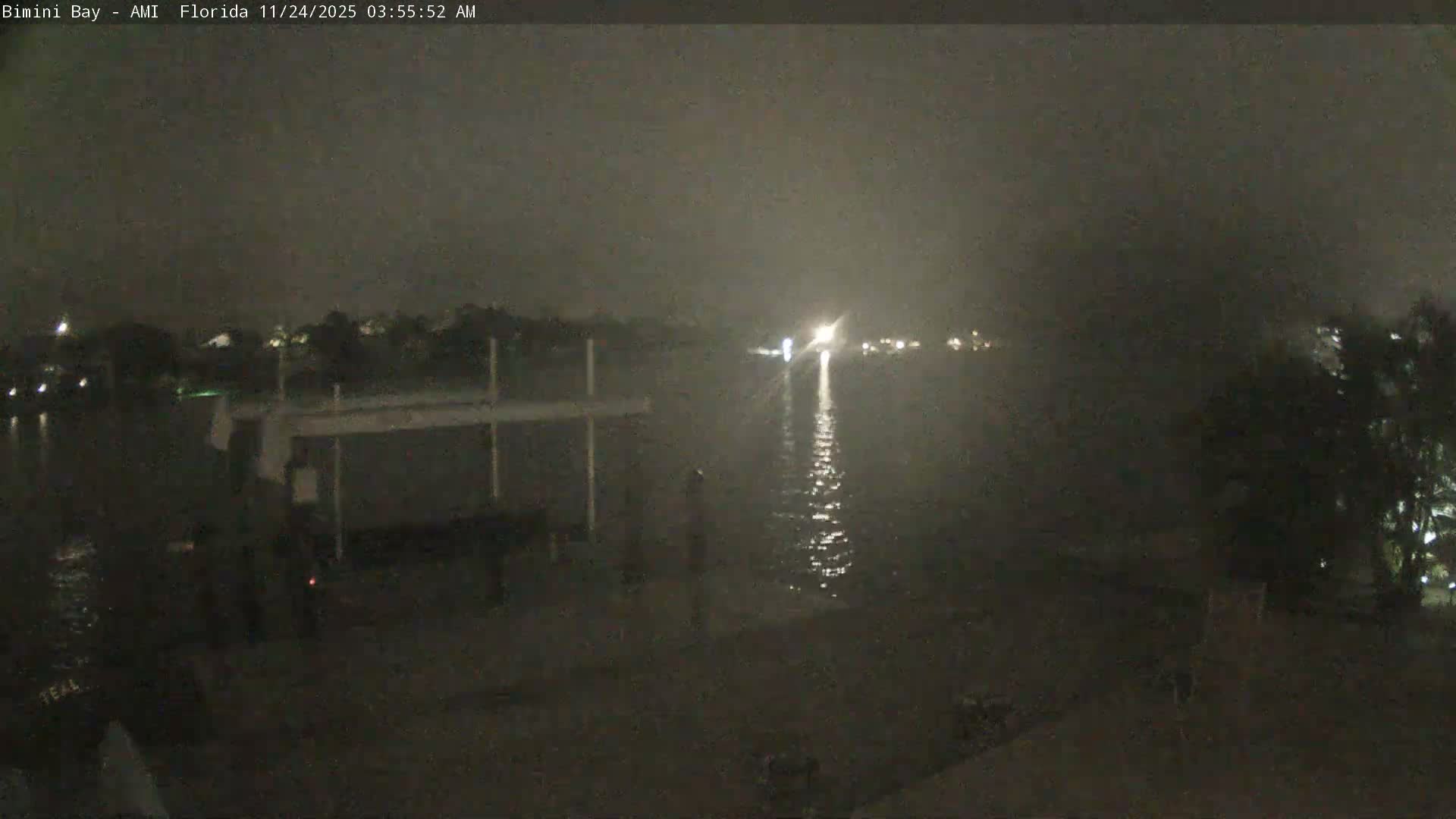 A very dark and foggy early morning view of a calm bay, with distant lights reflecting on the water and a dock partially visible on the left.