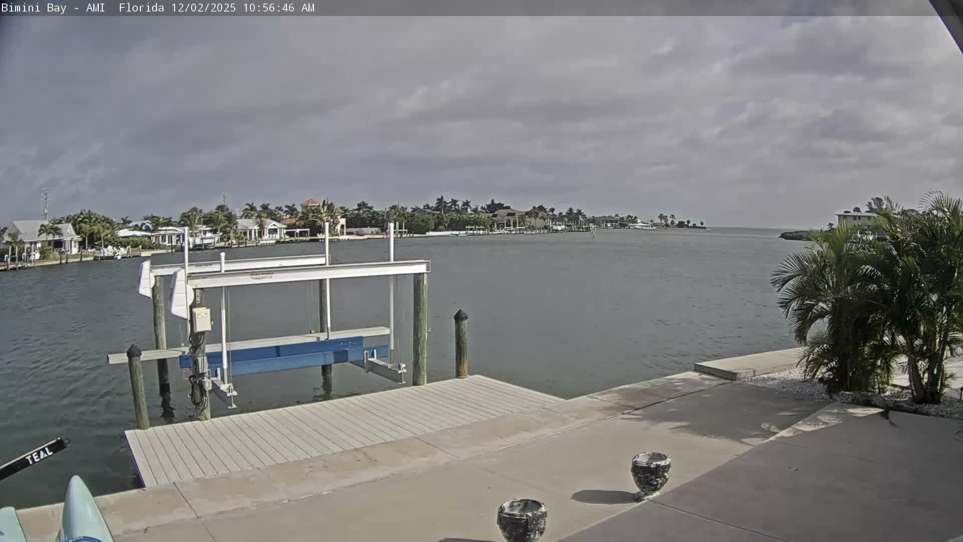 On a cloudy day, a view across a calm bay reveals a boat lift and dock in the foreground, with numerous waterfront homes nestled among palm trees lining the distant shore.
