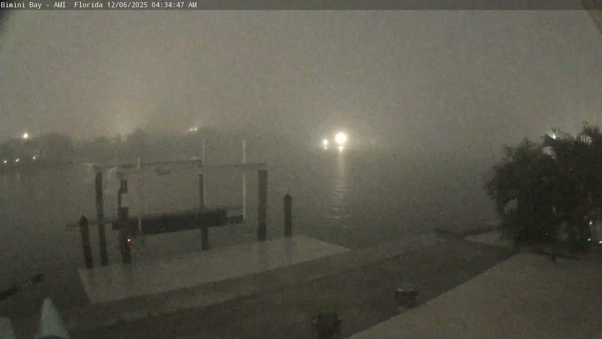 A boat dock stands on a dark, foggy bay or canal, with several bright lights piercing the thick mist in the distance.