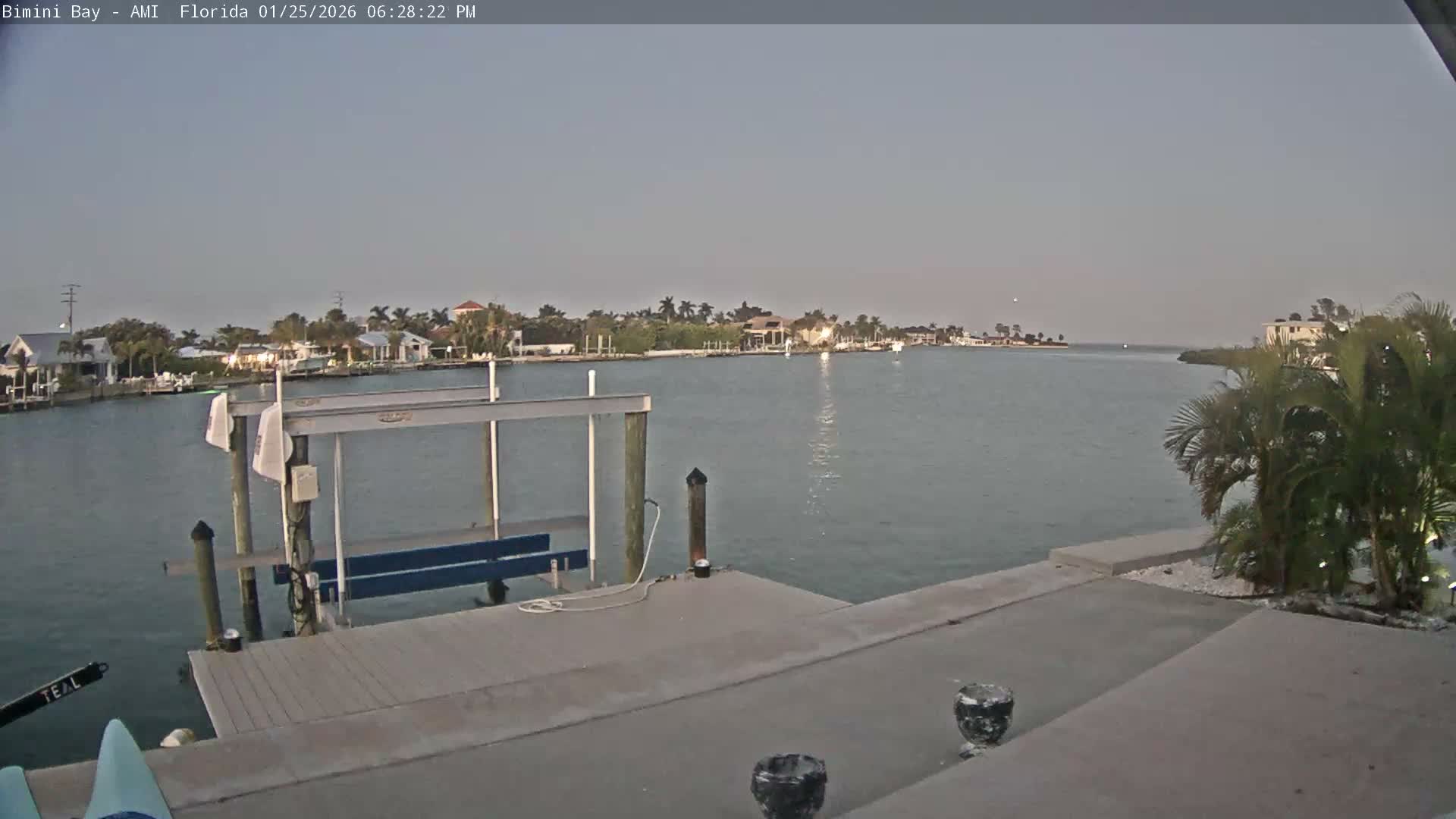 A calm and clear evening view captures a tranquil bay with waterfront homes and lush palm trees lining the distant shore, while a concrete dock and boat lift occupy the foreground.