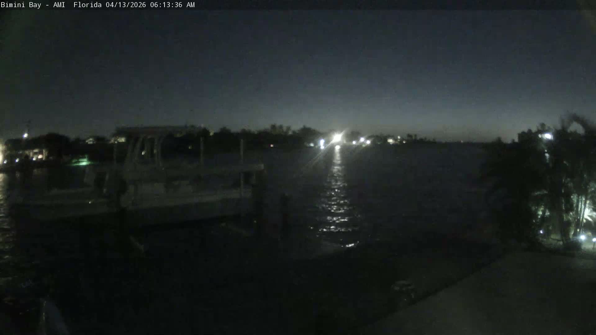 A calm and clear evening view captures a tranquil bay with waterfront homes and lush palm trees lining the distant shore, while a concrete dock and boat lift occupy the foreground.
