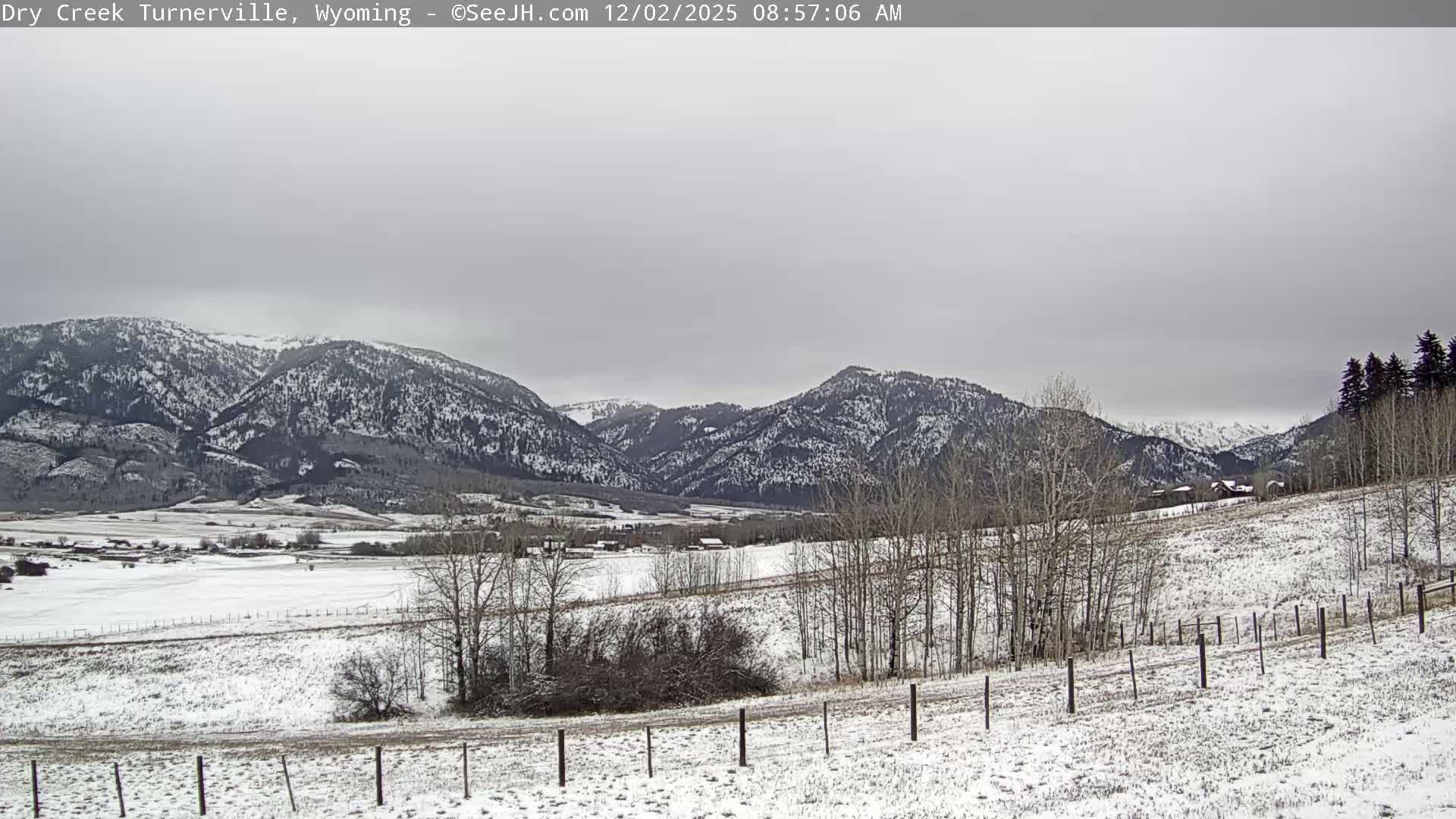 A wide panoramic view reveals a snow-covered valley with bare trees and distant farm buildings, backed by vast, snow-dusted mountains under a gray, overcast sky.