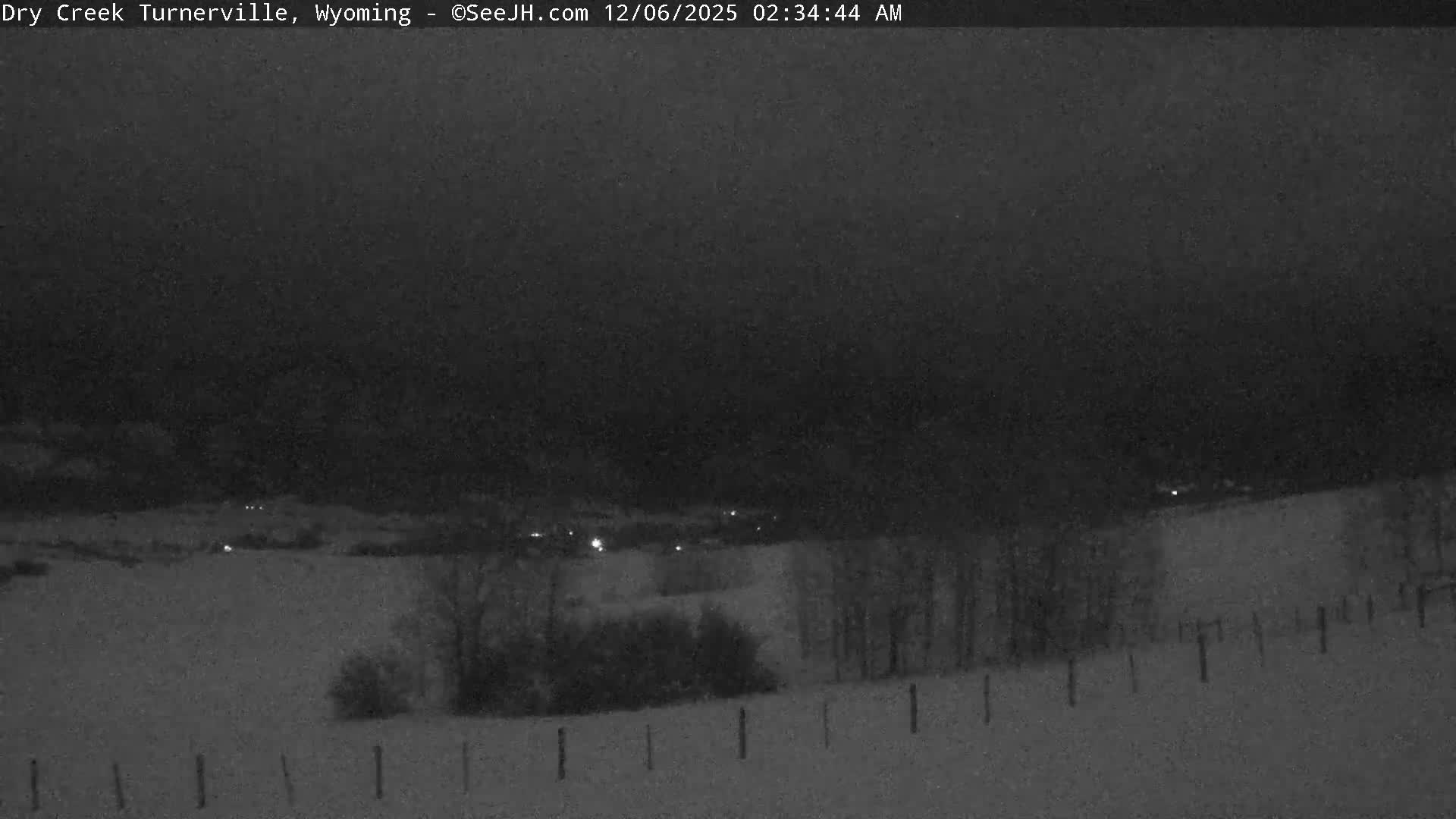 The image depicts a vast snow-covered landscape at night, featuring bare trees and distant scattered lights under a dark sky, indicating cold winter weather.