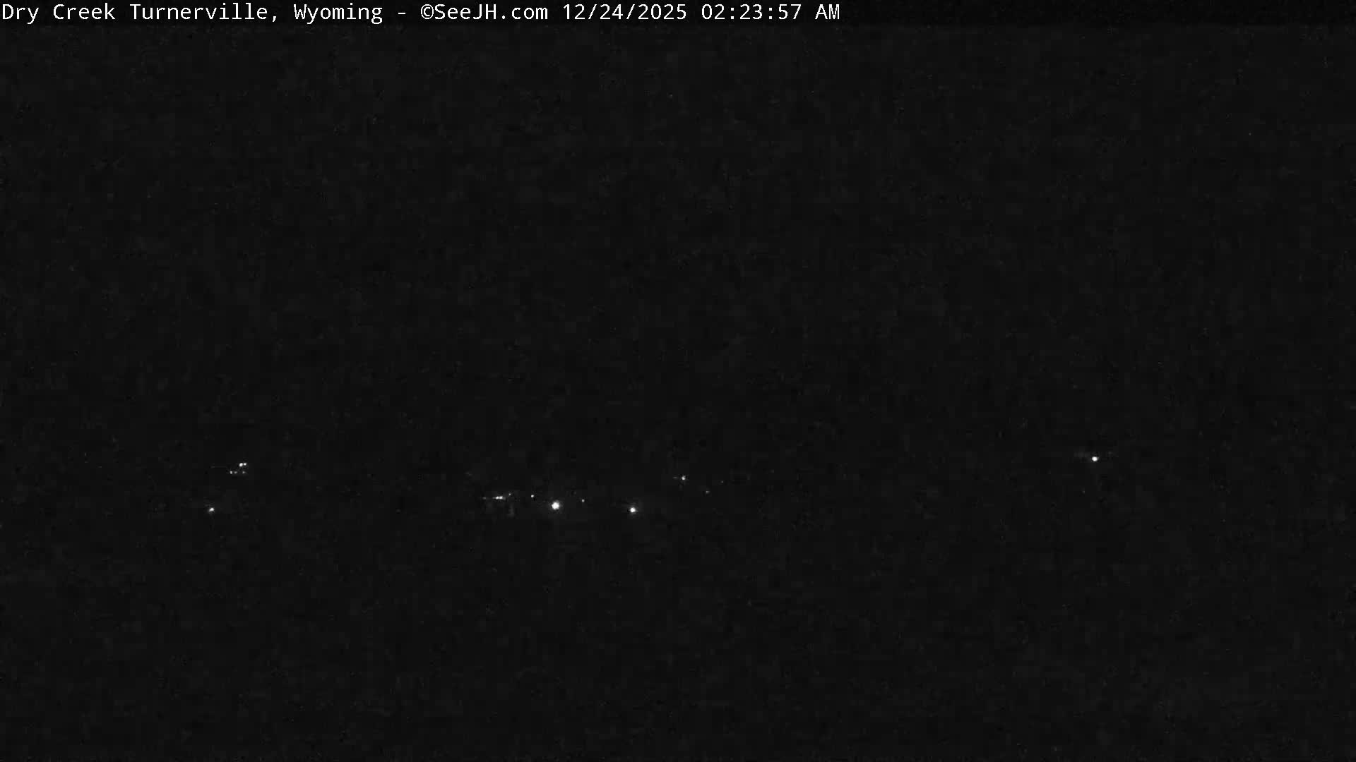 The image depicts a vast snow-covered landscape at night, featuring bare trees and distant scattered lights under a dark sky, indicating cold winter weather.