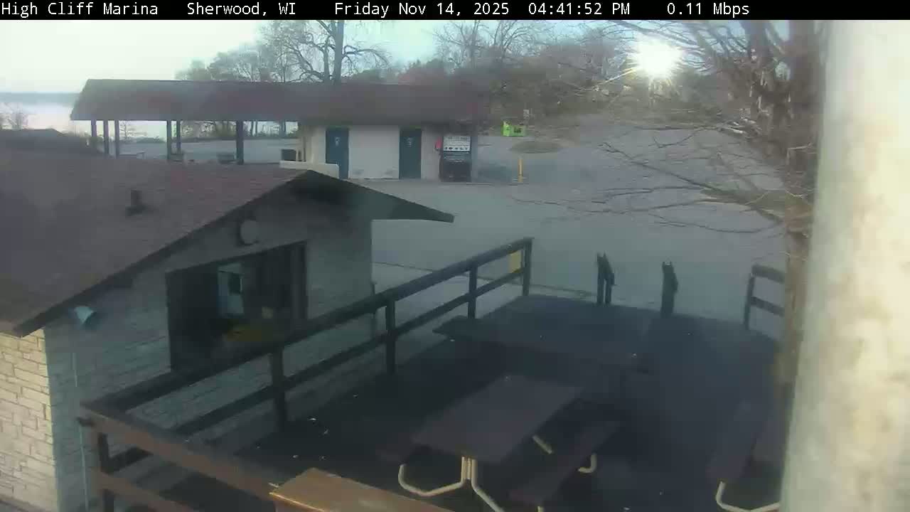 A hazy outdoor scene at a marina displays a building with a deck and picnic tables in the foreground, along with other structures and bare trees overlooking a body of water under a partially obscured bright sun.