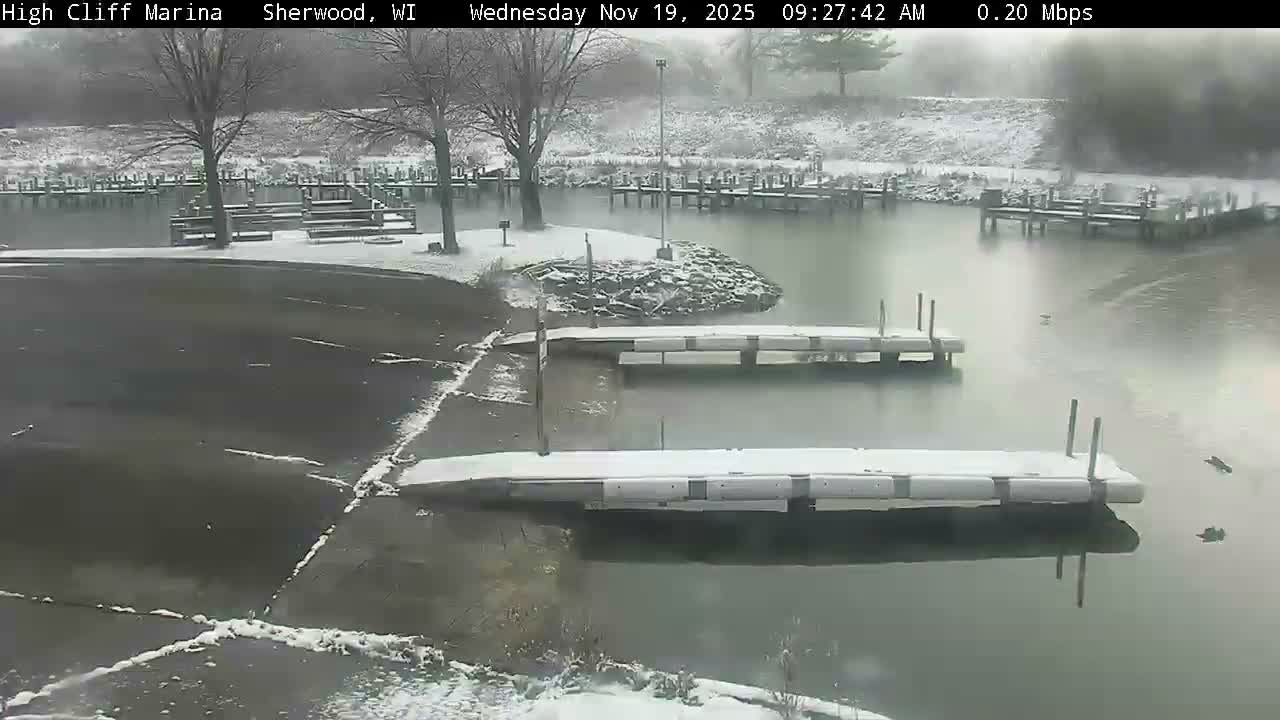 A nighttime grayscale view shows a marina with two empty docks floating on calm water, illuminated by a bright light source on the left shore where trees are visible, under clear conditions.