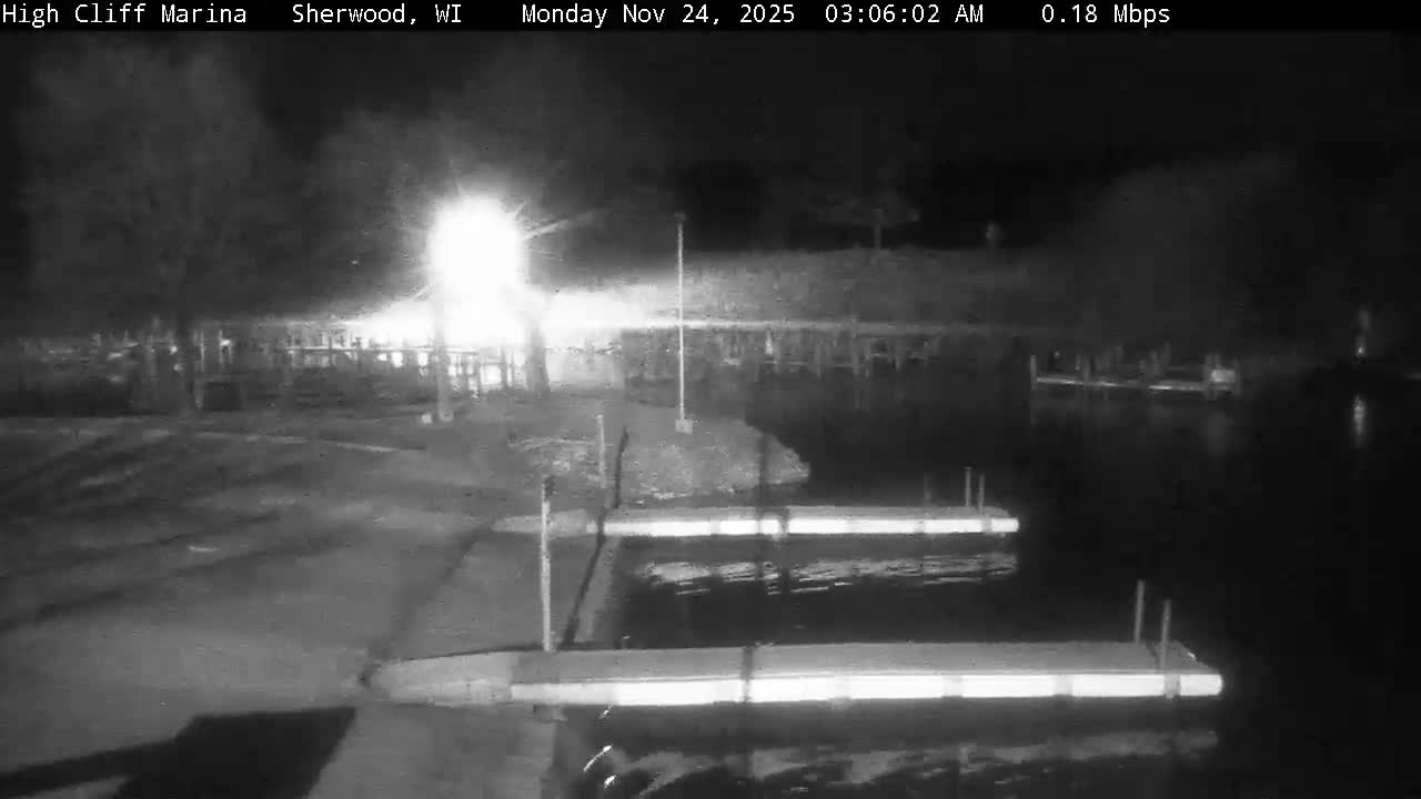 It is a clear, dark night at a marina, with two brightly lit docks extending into calm water in the foreground and a strong light source illuminating trees and other structures on the distant shore.