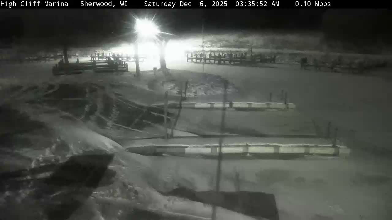 A snow-covered marina at night is brightly illuminated by a single light source, revealing docks extending into what appears to be partially frozen water under clear, cold conditions.