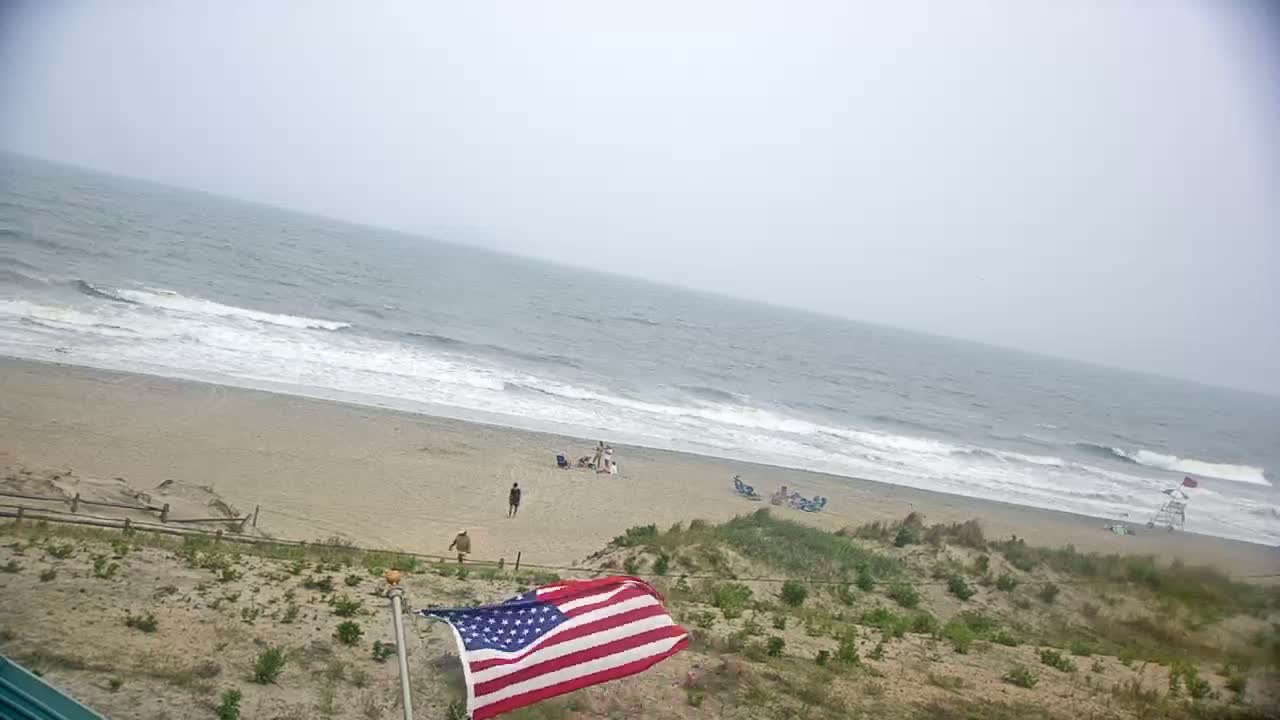 An American flag waves in the breeze above a beach scene with several people visible, under an overcast sky with moderate waves rolling onto the shore.
