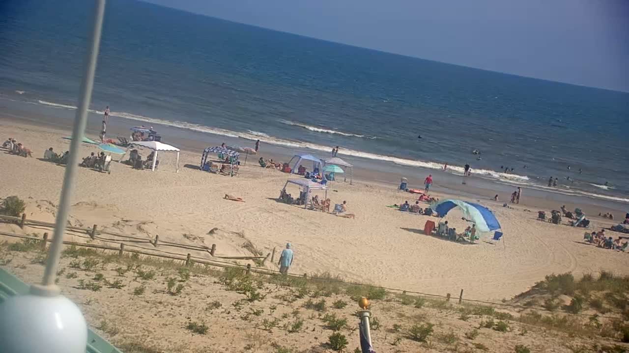 A sandy beach with people swimming and relaxing under umbrellas on a sunny day, viewed from a dune.