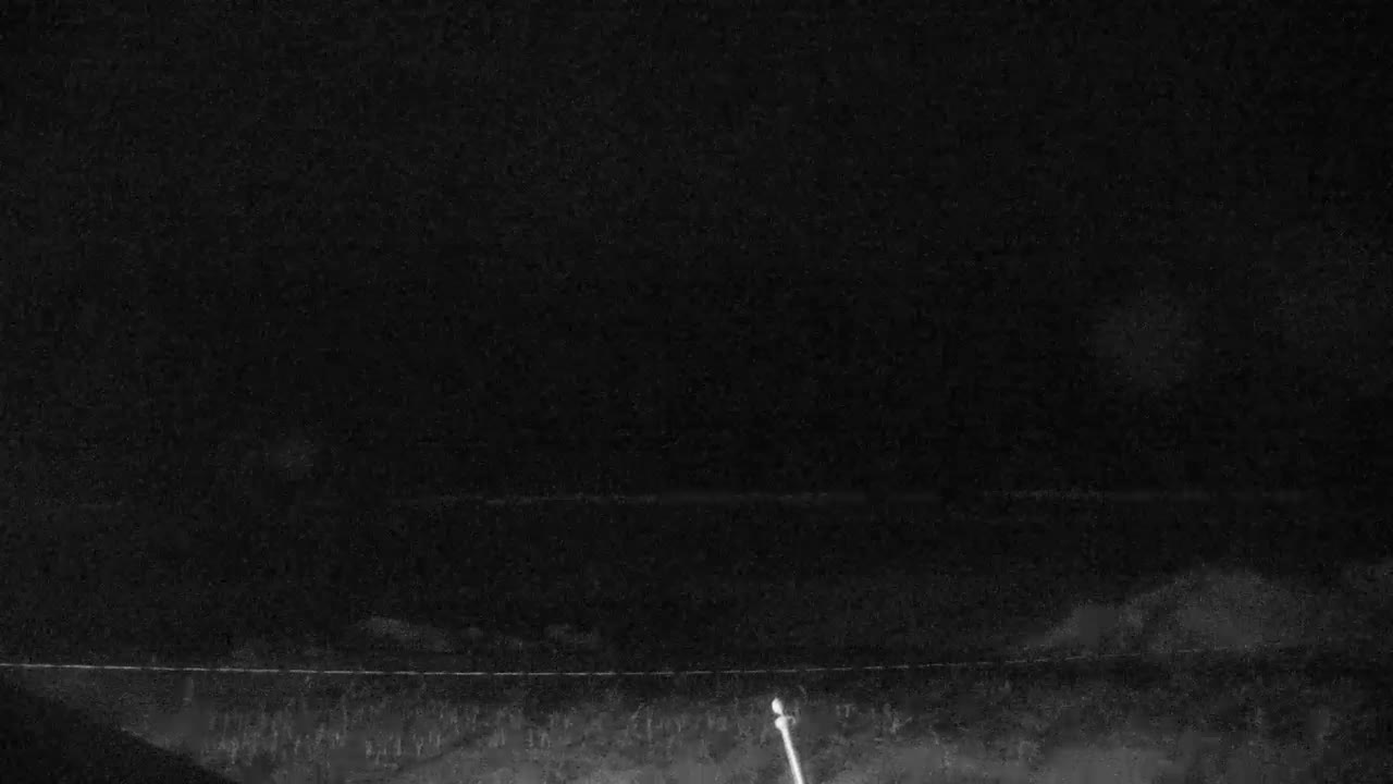 A dark, grainy outdoor night scene displays a faint horizontal line separating a dark, possibly overcast sky from indistinct ground below, featuring a light-colored pole in the lower right.