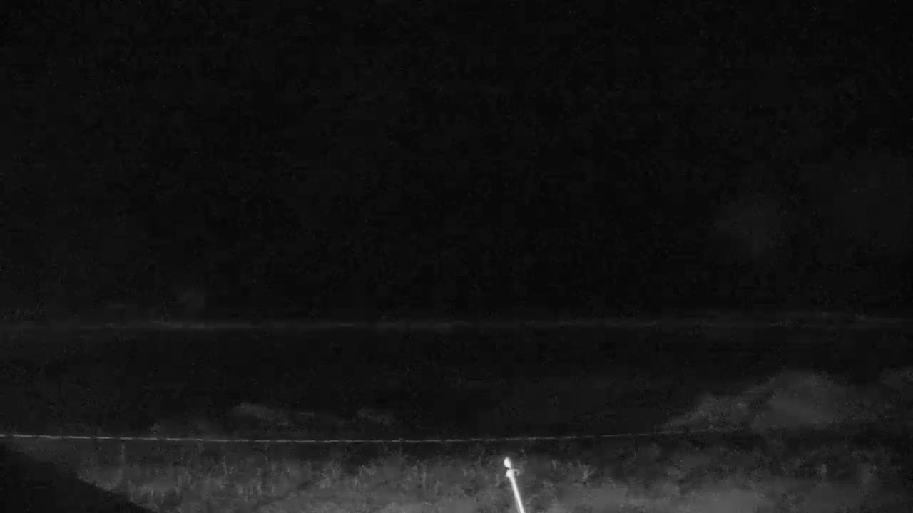 A very dark, grainy monochrome outdoor image, captured on a clear night, reveals a faint horizontal line across the middle distance, above a subtly textured foreground, with a distinct glowing marker in the bottom right.