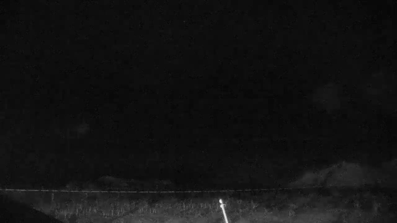 A dark, grainy image taken at night shows a subtle horizon line above a dimly discernible foreground with hints of vegetation and a bright light pole in the lower right, all beneath a faintly cloudy sky.
