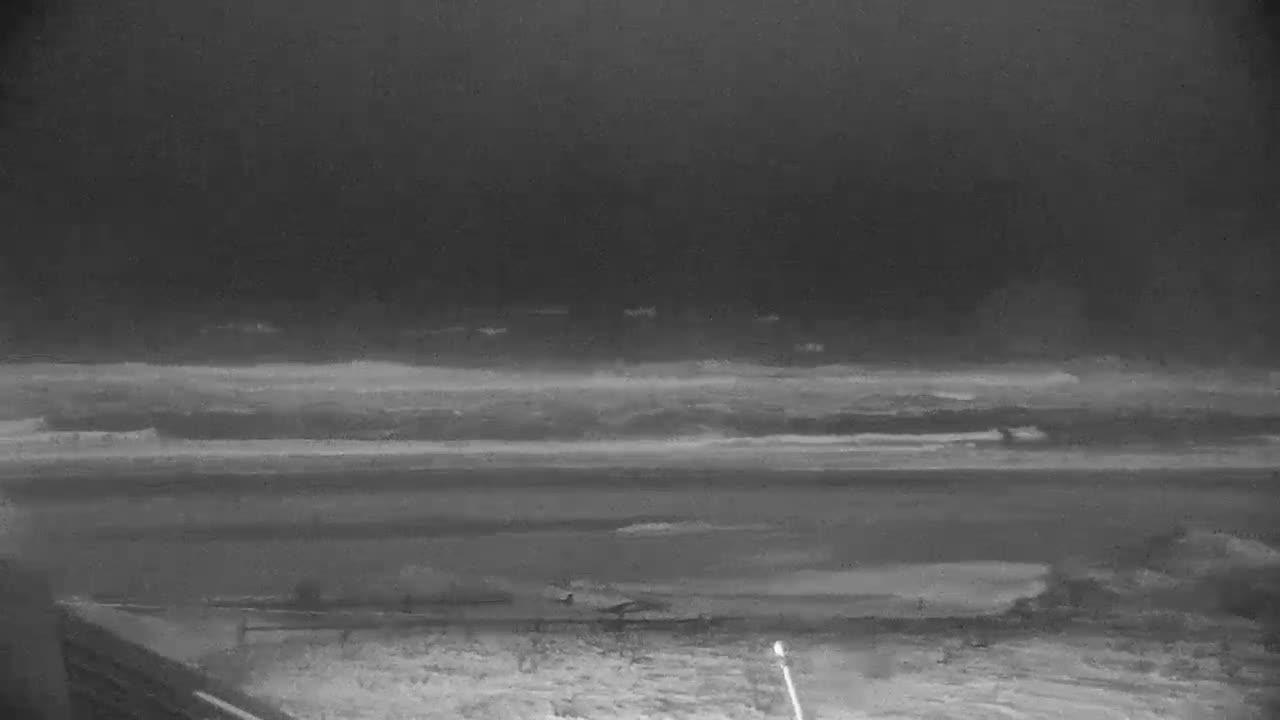 A dark, heavily overcast night reveals a vast, snow-covered or icy landscape, possibly an airfield, with a hazy horizon obscured by low-hanging clouds and a faint light source in the bottom right foreground.