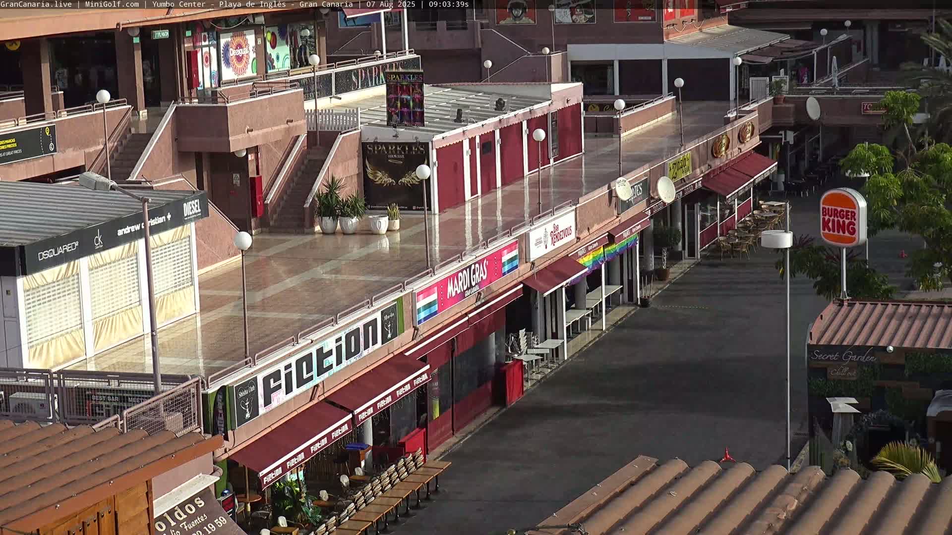 An outdoor view of a plaza with several shops and restaurants, some wet from a recent rain or early morning dew.