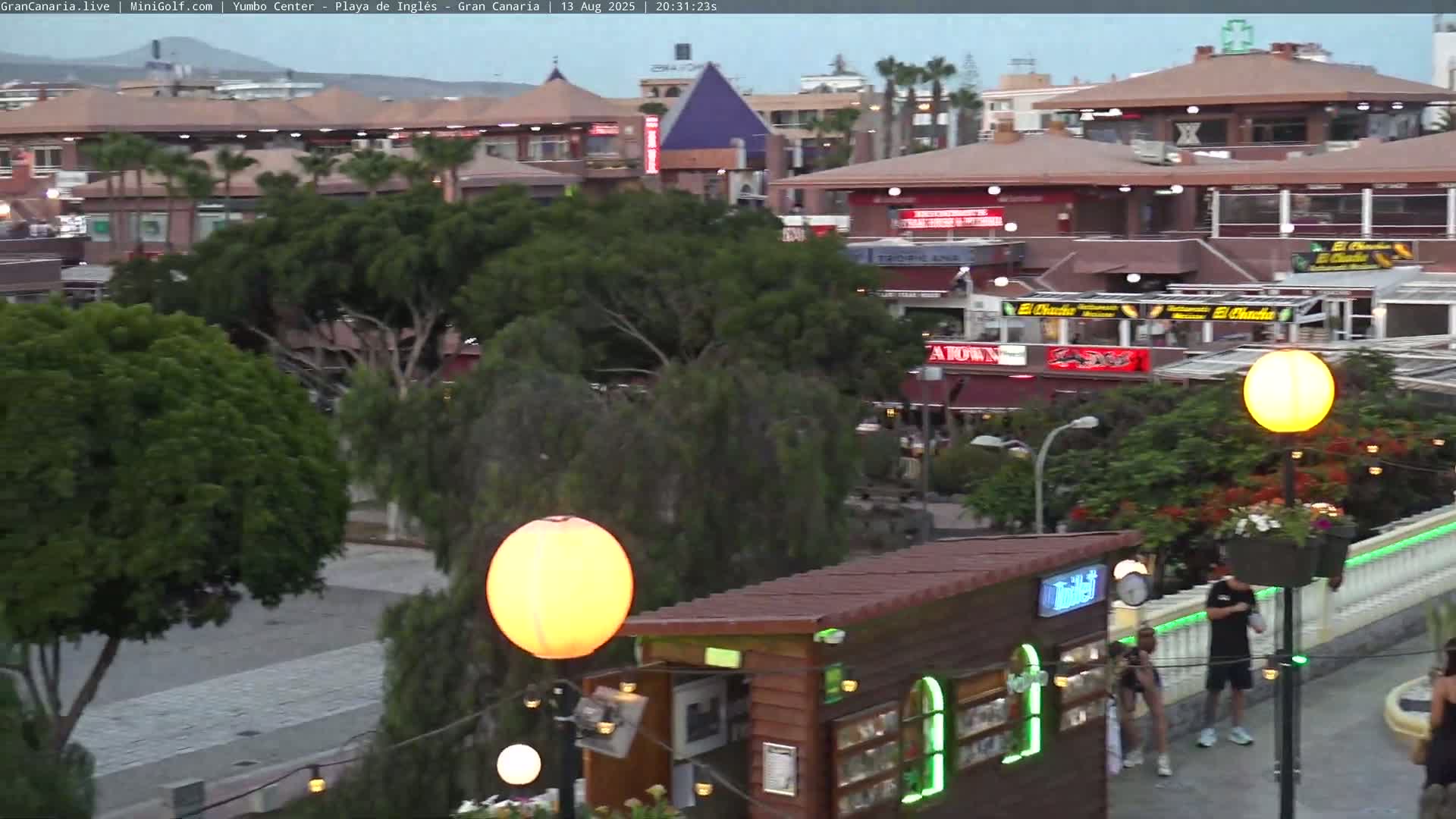 An outdoor evening scene shows a plaza with several restaurants and shops, illuminated by streetlights, with some pedestrians walking around under a twilight sky.