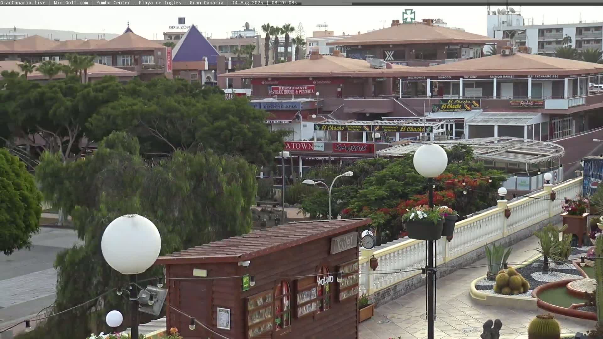 An outdoor scene shows a plaza with restaurants and shops, lush green trees, decorative landscaping, and white globe lights under a partly cloudy sky.