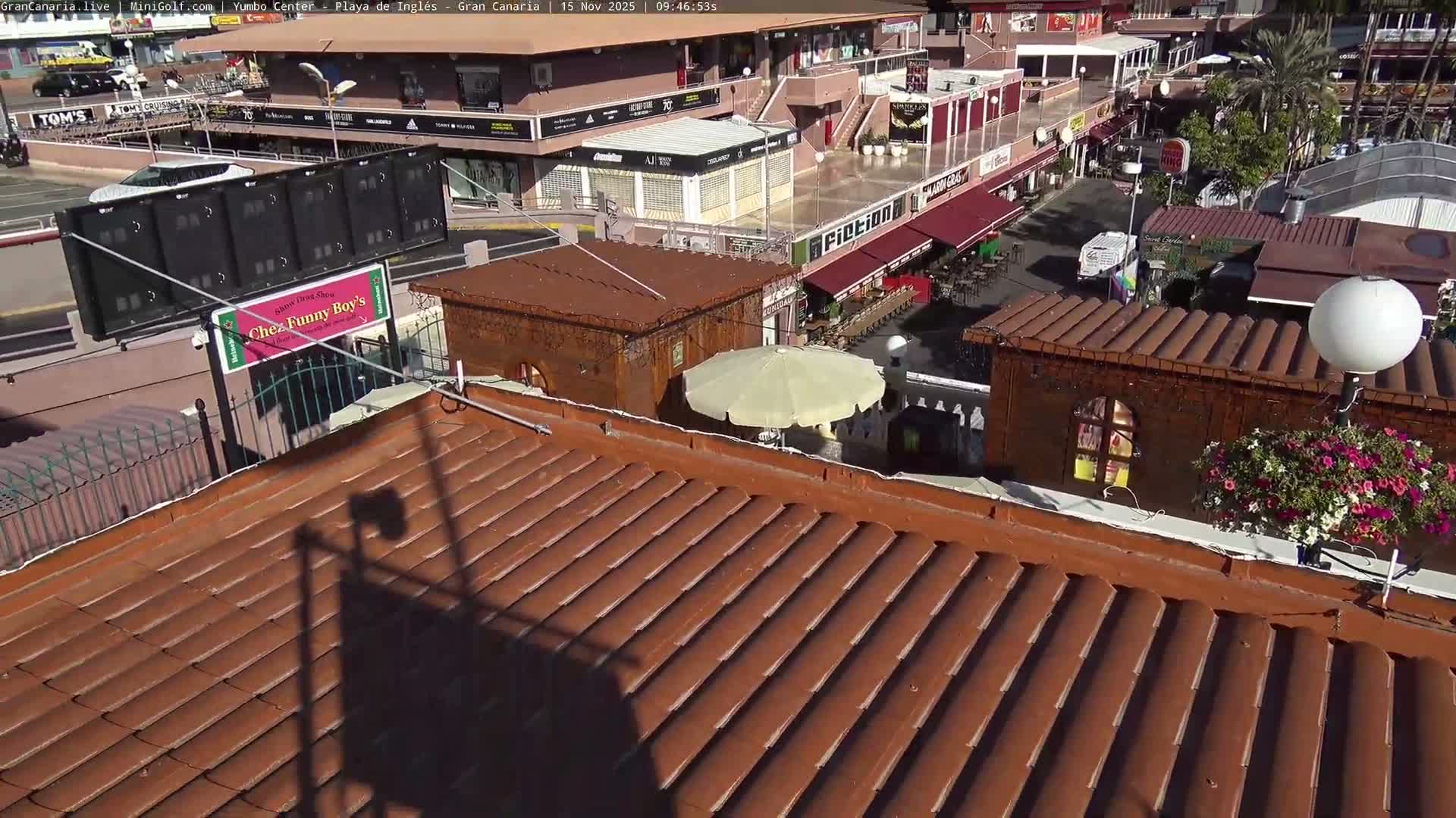 An elevated view under clear, sunny skies depicts a commercial plaza featuring shops with red awnings, outdoor seating, a street with a white van, and a large white umbrella, all seen from above a terracotta-tiled roof.