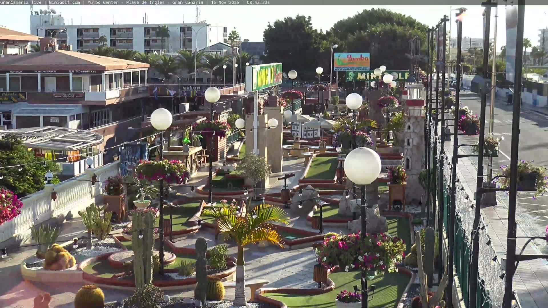 A sunny outdoor mini-golf course featuring numerous green fairways, diverse plants including cacti and flowering bushes, decorative white spherical lights, and a miniature lighthouse, with multi-story buildings and a street visible in the background.
