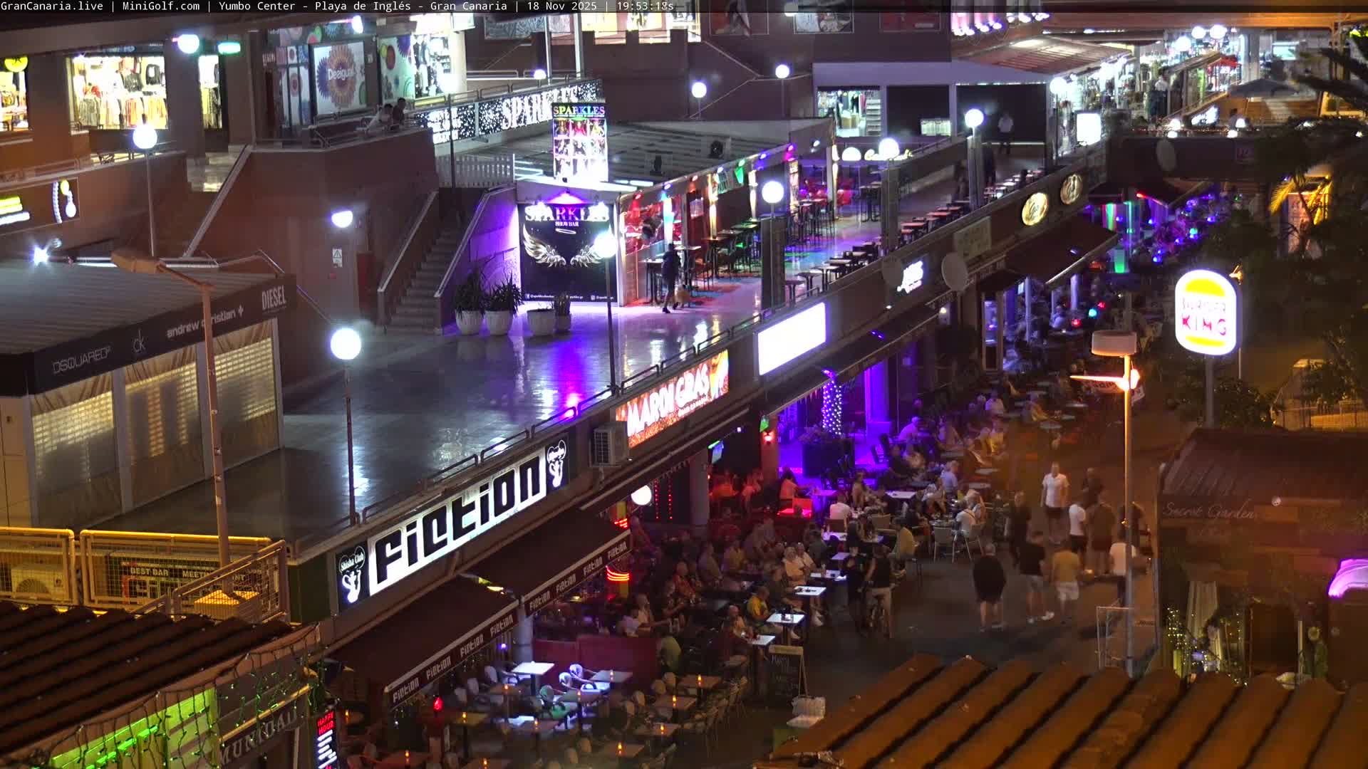 A sunny outdoor mini-golf course featuring numerous green fairways, diverse plants including cacti and flowering bushes, decorative white spherical lights, and a miniature lighthouse, with multi-story buildings and a street visible in the background.