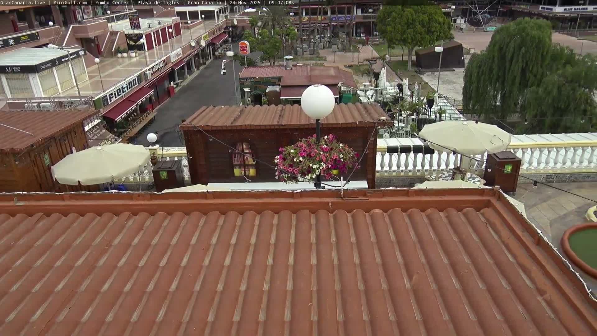 An elevated view on a clear, bright day shows a reddish-brown tiled roof in the foreground, leading to a wooden structure adorned with a floral lamp post, and an outdoor multi-level shopping complex with various shops, walkways, and green spaces in the background.