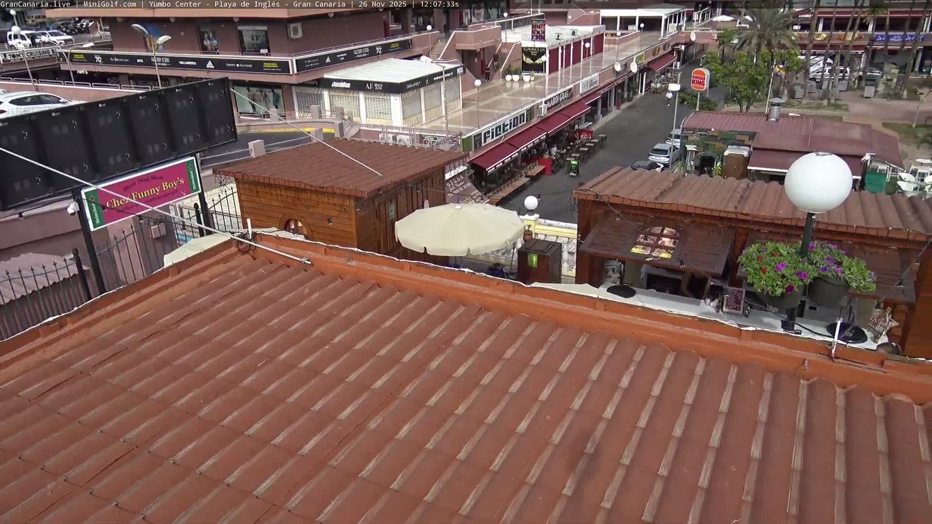 An elevated view on a clear, bright day shows a reddish-brown tiled roof in the foreground, leading to a wooden structure adorned with a floral lamp post, and an outdoor multi-level shopping complex with various shops, walkways, and green spaces in the background.