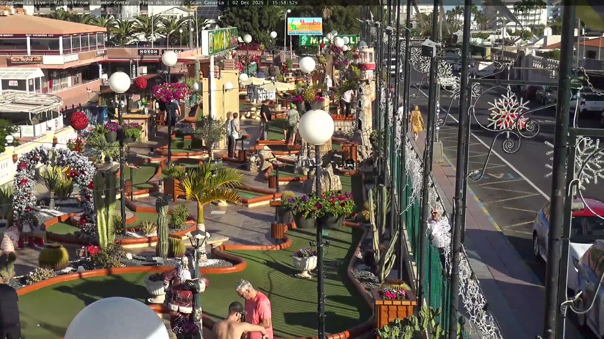 A sunny outdoor miniature golf course, decorated with festive holiday lights, garlands, palm trees, and cacti, hosts several people playing golf while others stroll on a neighboring street.