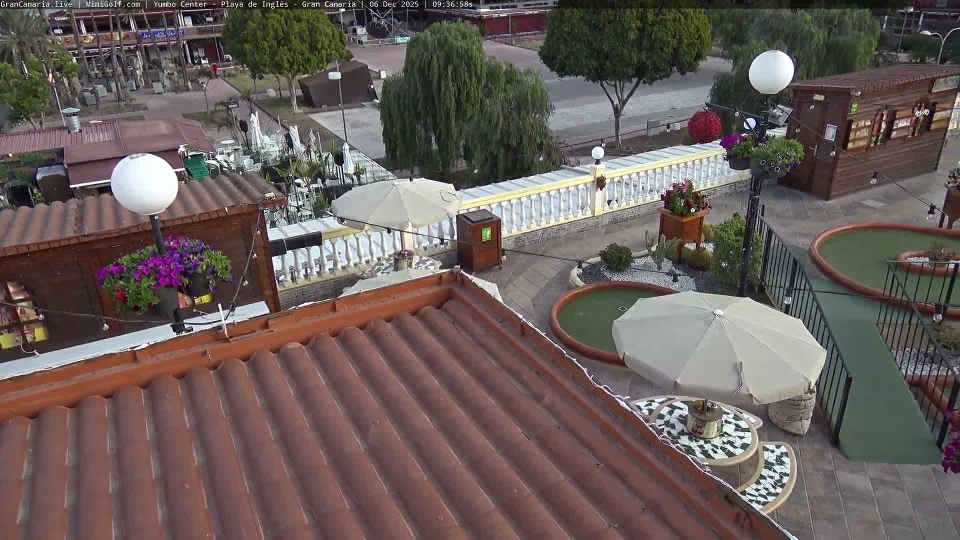 An elevated view reveals an outdoor mini-golf course with two green holes, various patio seating areas with beige umbrellas, numerous potted purple and red flowers, a white balustrade, and distant buildings, all under bright, clear skies.