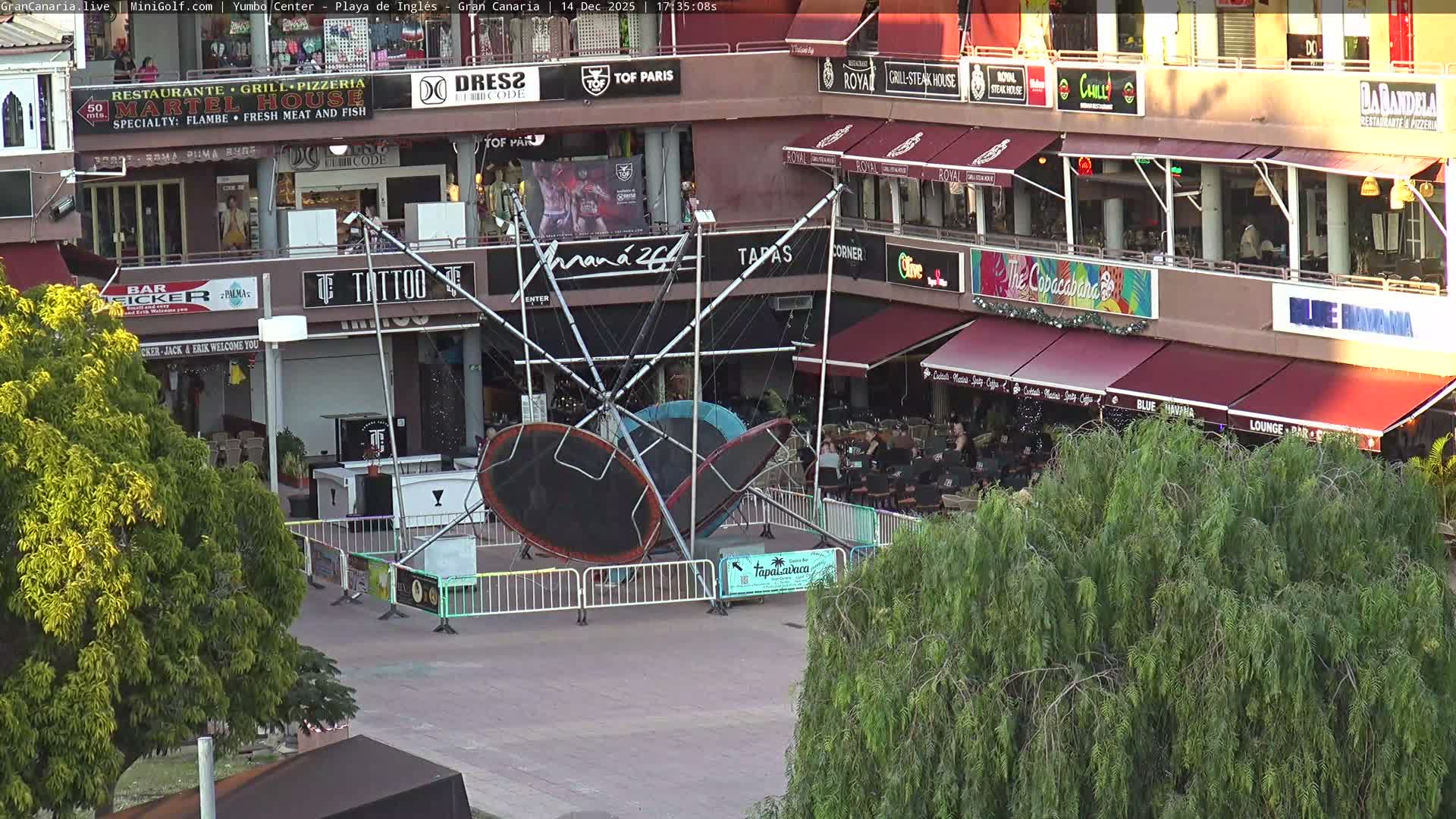 An elevated view reveals an outdoor mini-golf course with two green holes, various patio seating areas with beige umbrellas, numerous potted purple and red flowers, a white balustrade, and distant buildings, all under bright, clear skies.