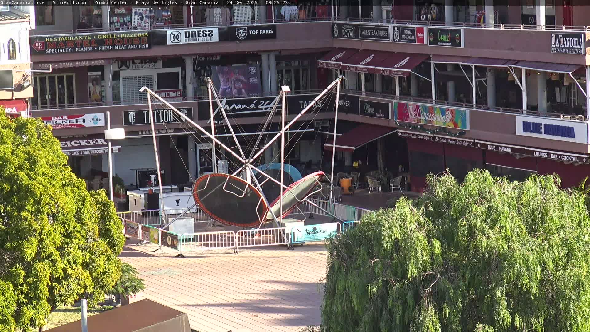 An elevated view reveals an outdoor mini-golf course with two green holes, various patio seating areas with beige umbrellas, numerous potted purple and red flowers, a white balustrade, and distant buildings, all under bright, clear skies.