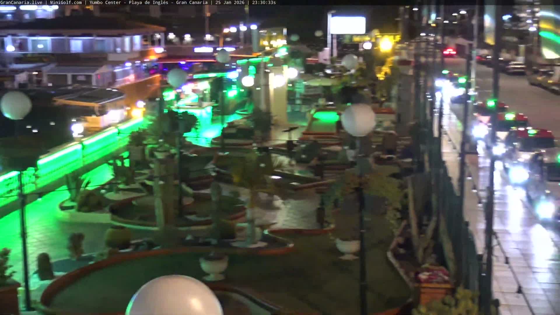 Vibrant green lighting and white spherical lanterns illuminate an outdoor mini-golf course at night, with blurred buildings and a street with cars visible in the background under clear skies.