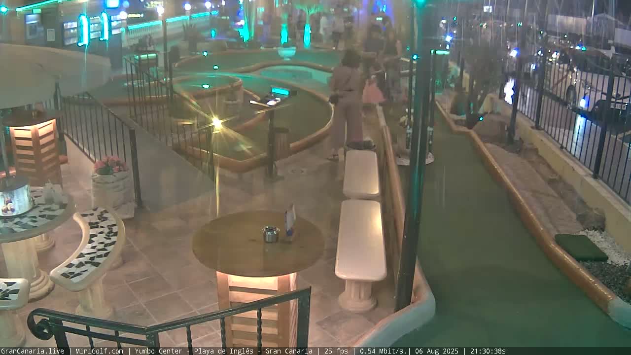 An outdoor, nighttime scene shows a mini-golf course with people playing, and nearby seating areas with small tables.
