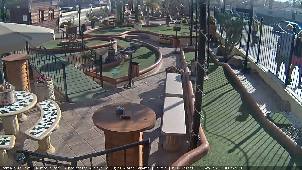 A brightly sunlit outdoor miniature golf course features winding green fairways, paved paths, a patio with mosaic tables and benches, and a few pedestrians walking past a black metal fence on the right.