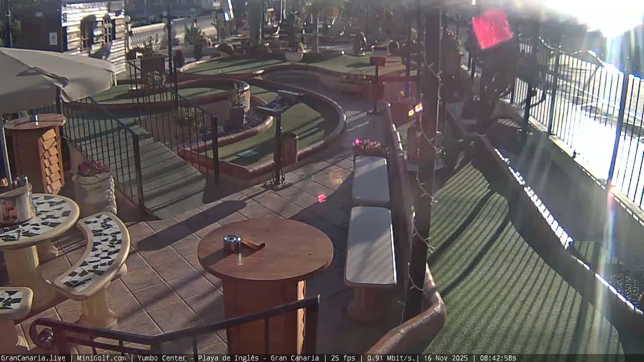 The image shows an outdoor mini-golf course with tiled patio seating areas, tables, and benches under a bright, sunny sky.