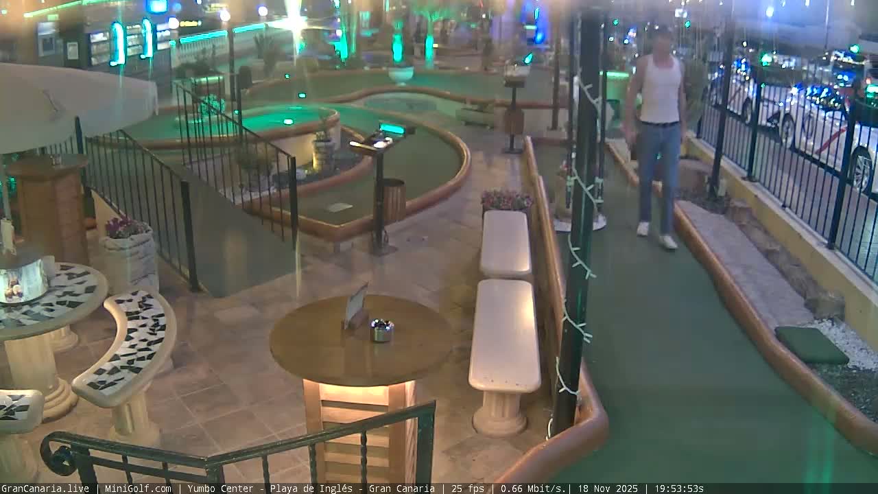 The image shows an outdoor mini-golf course with tiled patio seating areas, tables, and benches under a bright, sunny sky.