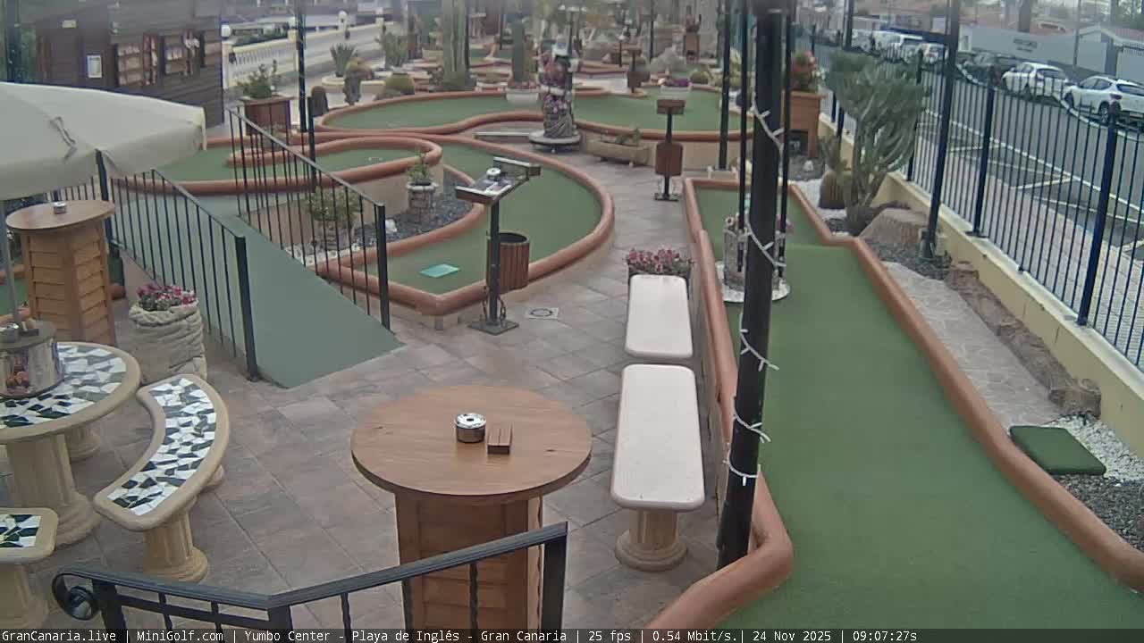 The image depicts an outdoor mini-golf course with green lanes, various obstacles, multiple seating areas with tables and benches, and a street with parked cars visible beyond a fence, all under an overcast sky.