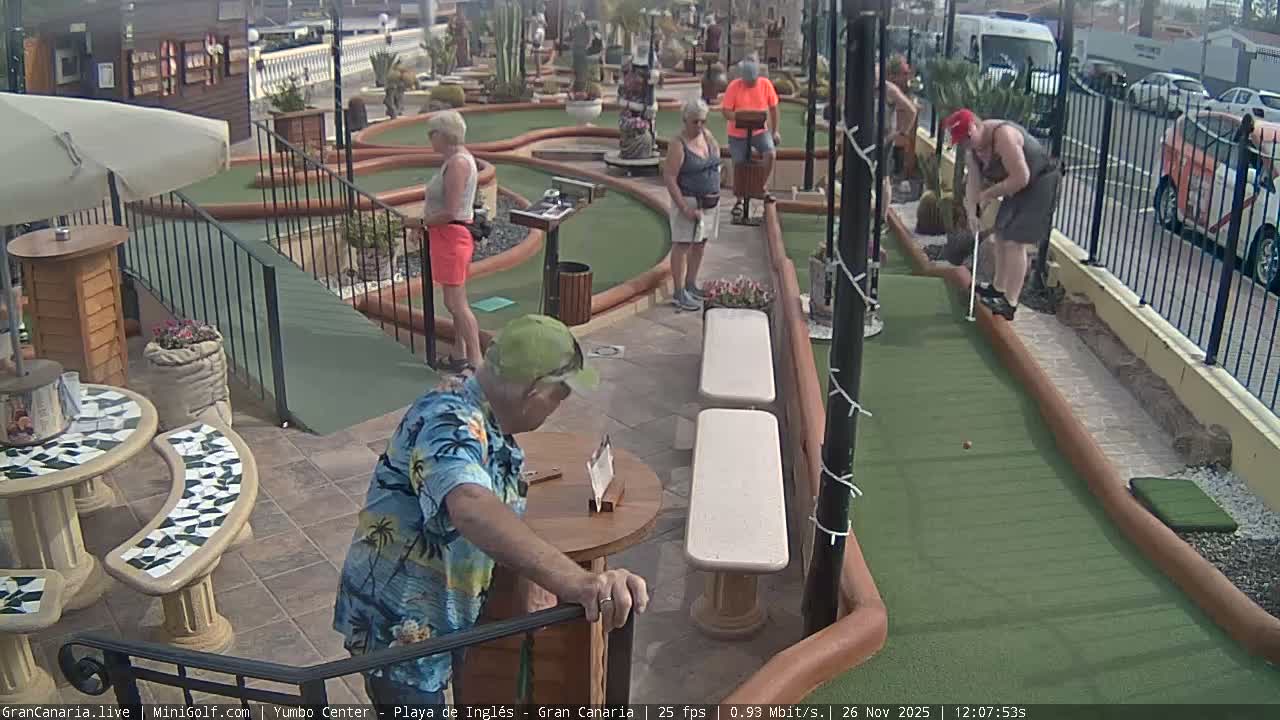 The image depicts an outdoor mini-golf course with green lanes, various obstacles, multiple seating areas with tables and benches, and a street with parked cars visible beyond a fence, all under an overcast sky.