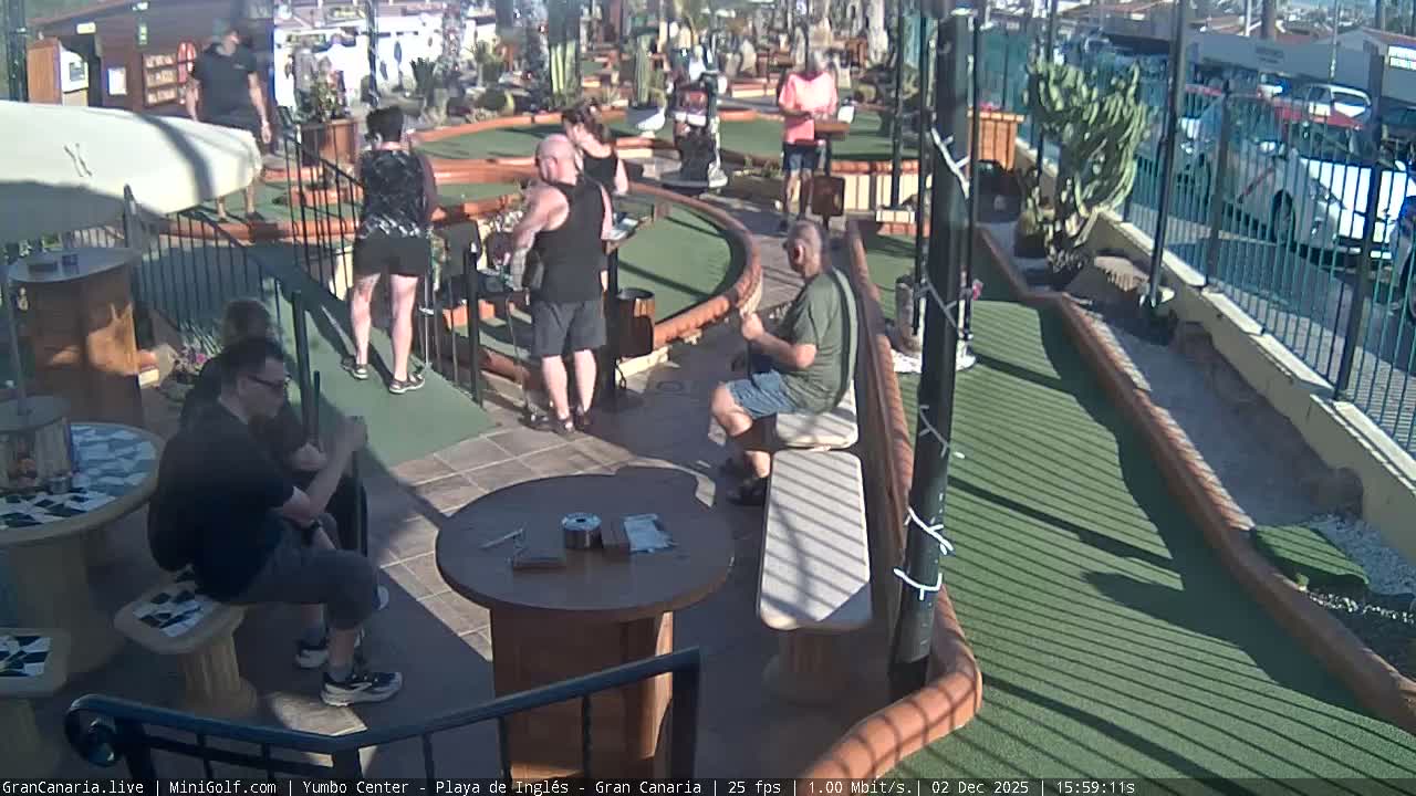 On a bright, sunny day, several people are seen playing or observing on an outdoor mini-golf course.
