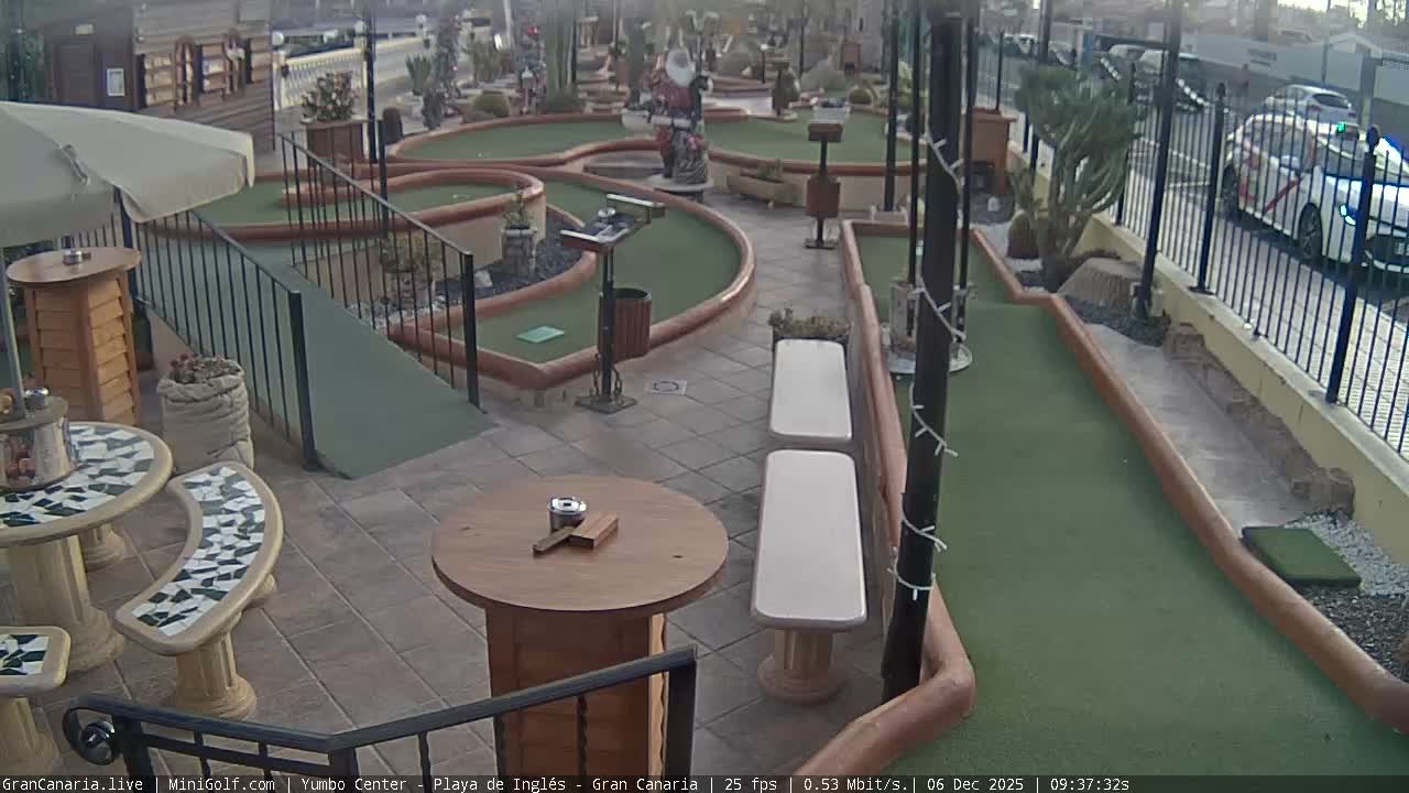 An outdoor miniature golf course with green fairways, obstacles including a Santa Claus statue, and adjacent patio seating areas with tables and benches, is seen under clear and mild morning conditions.