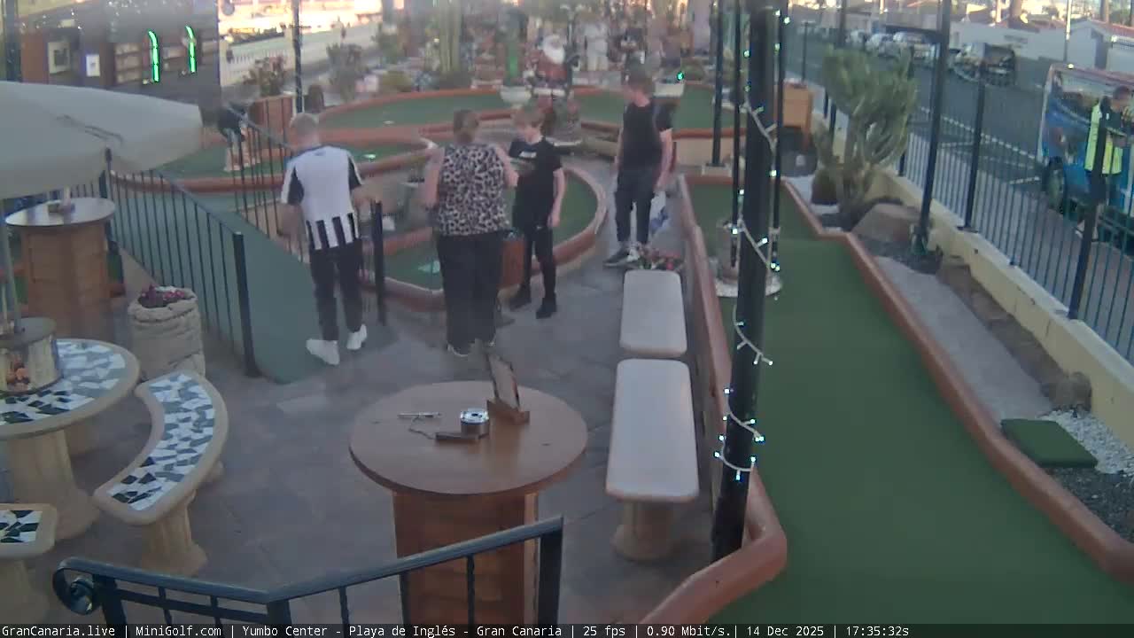 An outdoor miniature golf course with green fairways, obstacles including a Santa Claus statue, and adjacent patio seating areas with tables and benches, is seen under clear and mild morning conditions.
