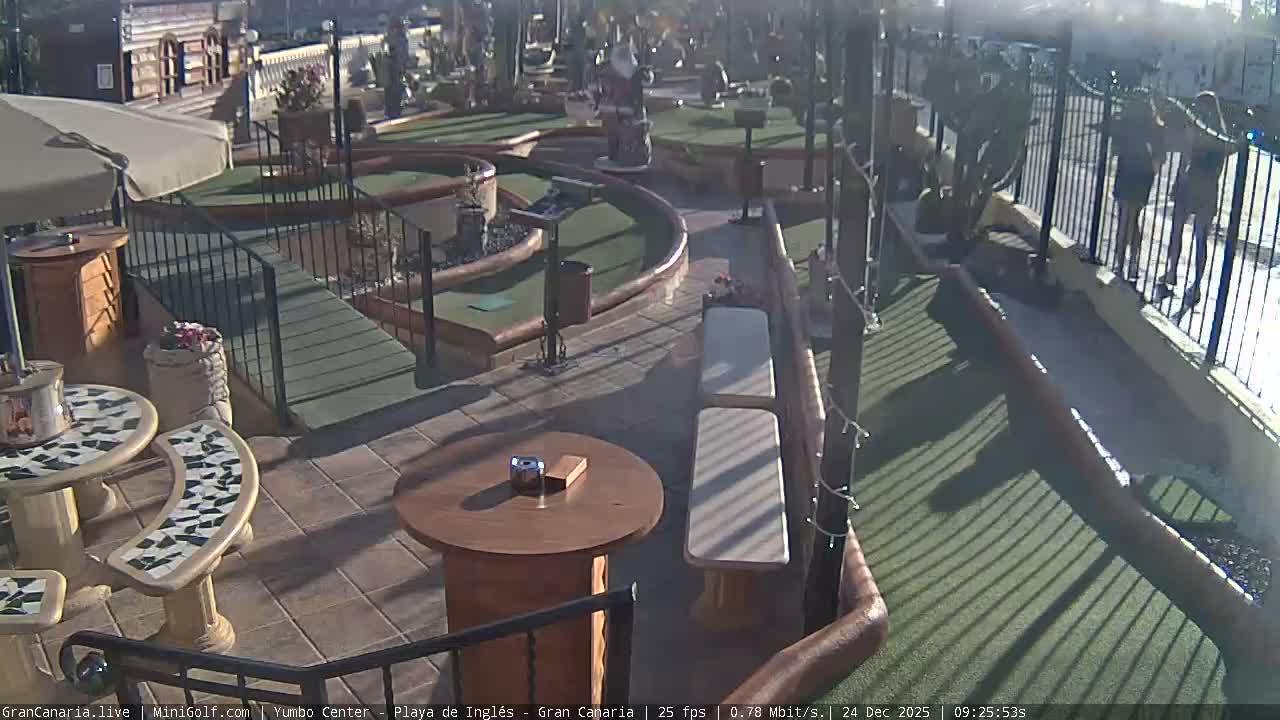 An outdoor miniature golf course with green fairways, obstacles including a Santa Claus statue, and adjacent patio seating areas with tables and benches, is seen under clear and mild morning conditions.